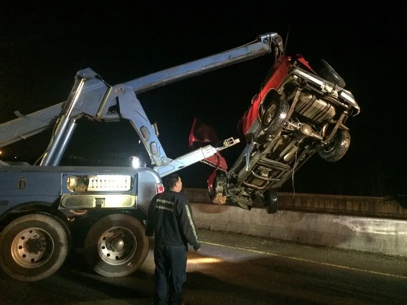 At night, a wrecker boom lifts a red pickup back over a concrete barrier as the operator watches.