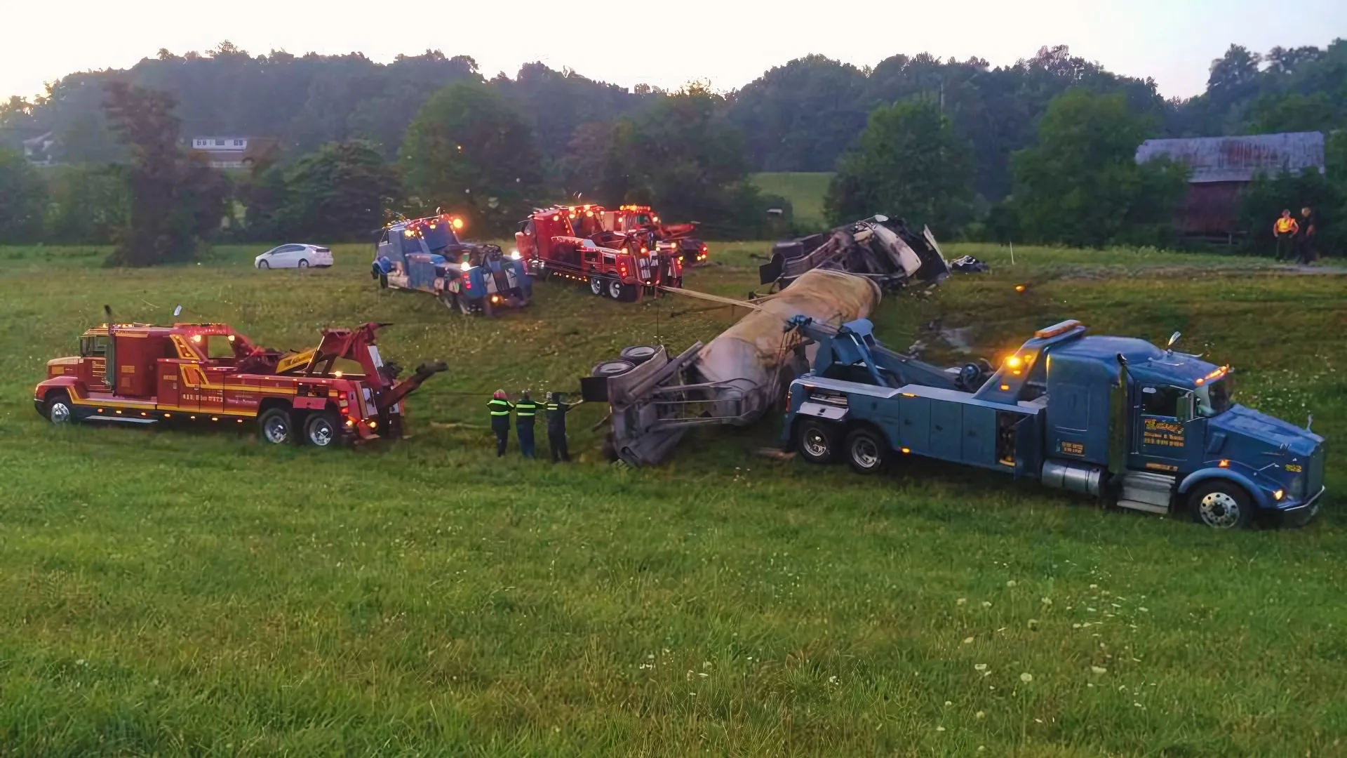 Several wreckers, blue and red, recovering an overturned trailer in a field at dusk while the crew works on foot.