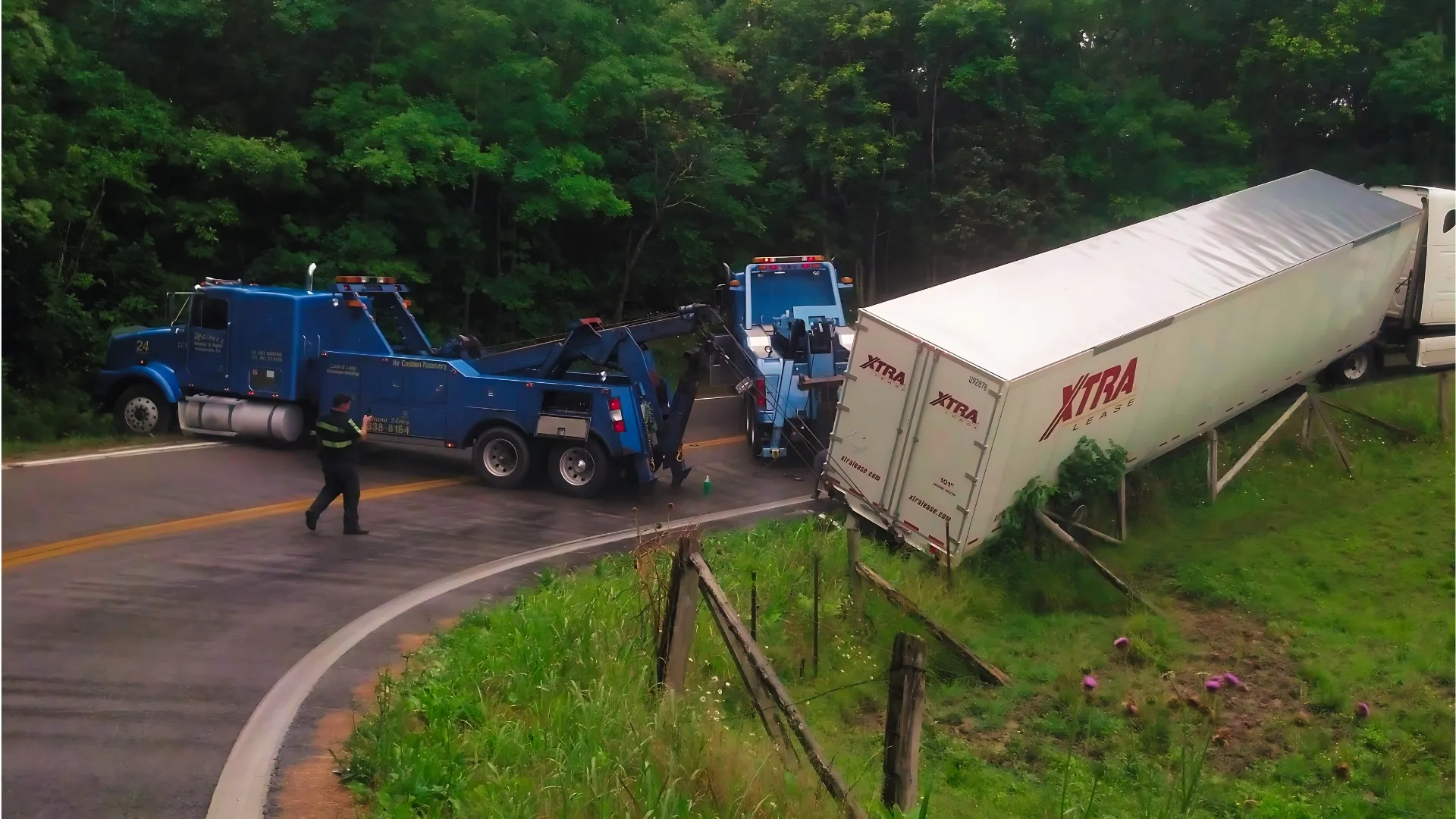 A blue Malone’s heavy wrecker on a curve, recovering a trailer that has gone off the road and down the bank.